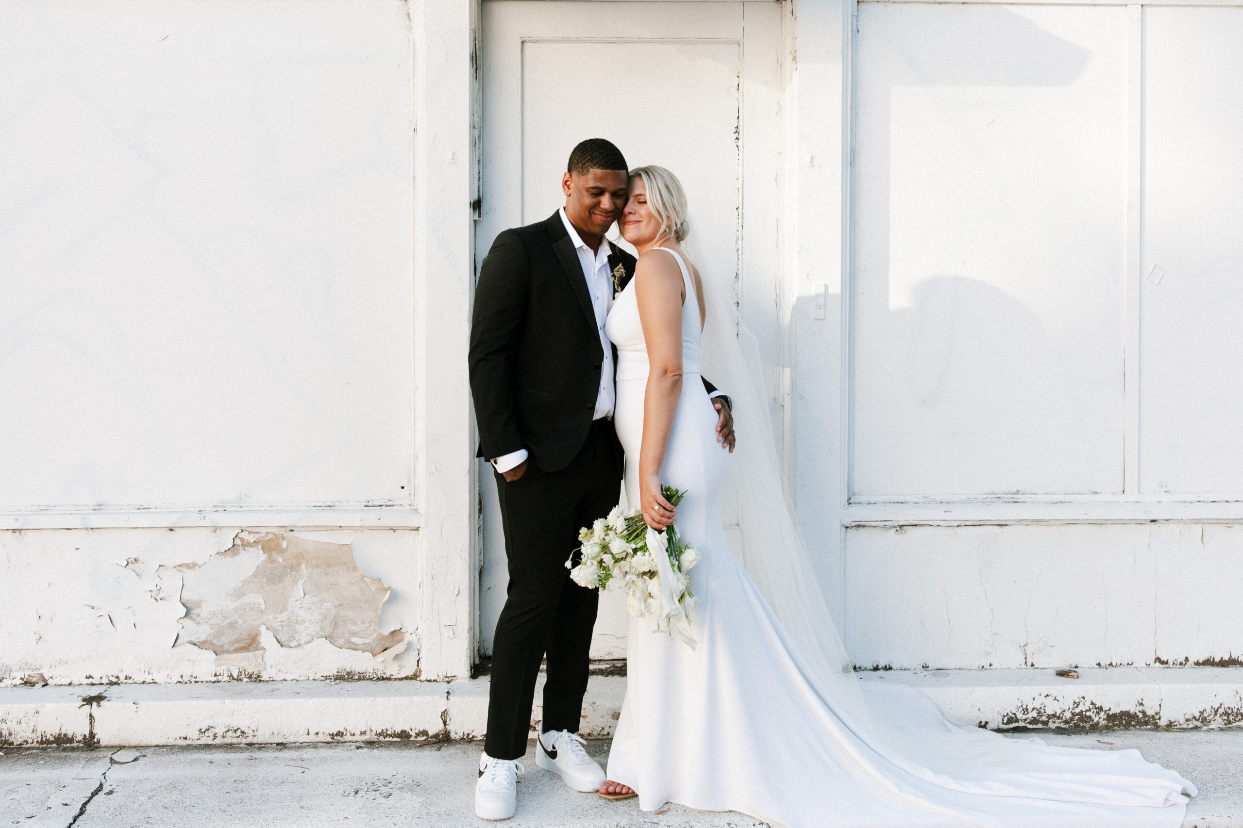 Bride and Groom Standing Against White Wall on Sidewalk